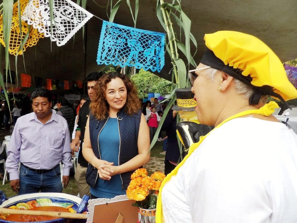 Catalina Verónica Atenco Pérez, Síndica Municipal de Cuernavaca, interactúa con ciudadanos durante un evento cultural con decoraciones tradicionales y gastronomía local.