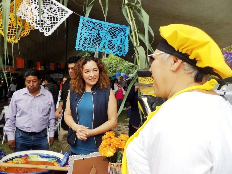 Catalina Verónica Atenco Pérez, Síndica Municipal de Cuernavaca, interactúa con ciudadanos durante un evento cultural con decoraciones tradicionales y gastronomía local.