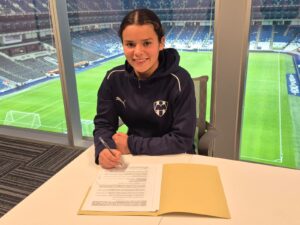 Isabella Lavín, Joven futbolista con sudadera del equipo Monterrey (Rayadas) firmando un documento que se entiende como su contrato en una oficina con vista al estadio BBVA. Sonríe mientras sostiene un bolígrafo, con una carpeta amarilla sobre la mesa.