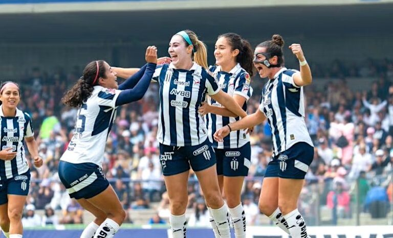 La imagen muestra a un grupo de jugadoras del equipo de fútbol femenil Rayadas de Monterrey celebrando en la cancha. Visten el uniforme del club, que consiste en franjas verticales azul marino y blanco, con detalles de patrocinadores como BBVA. Una de las jugadoras usa una máscara facial protectora, y todas tienen expresiones de alegría mientras se felicitan entre sí. Al fondo, se observa un estadio con público en las gradas, lo que indica que se trata de un partido con espectadores.